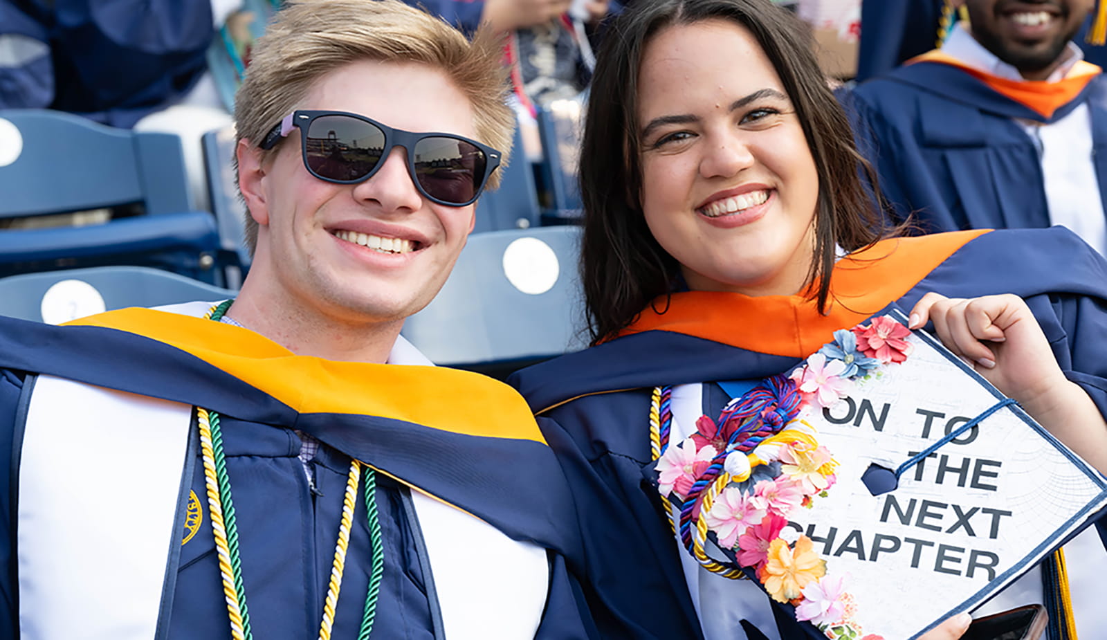 Two students in a crowd, with a woman holding a decorated cap with the words "onto the next chapter"